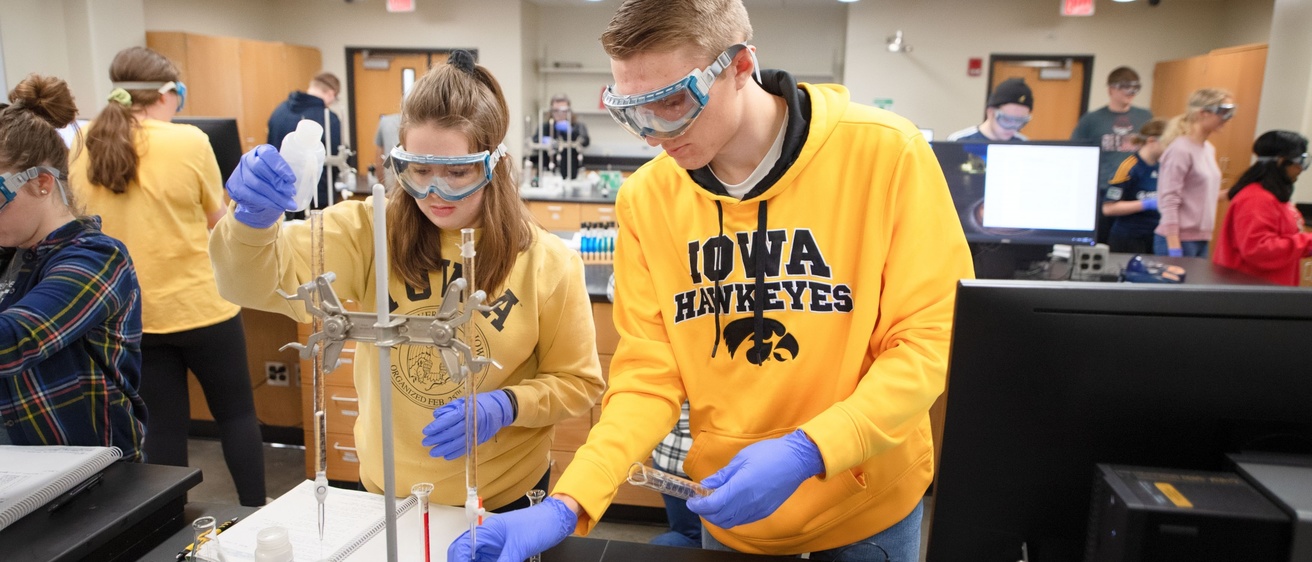 Two people with safety goggles working with some chemistry equipment